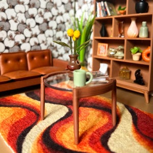 May include: A miniature living room scene featuring a brown sofa, a round glass coffee table with a light green mug, and a brown vase with yellow flowers. A wooden bookshelf displays various small decorative items, all set on a wavy orange rug.