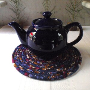 May include: A dark blue teapot with a lid sits on a round, colorful fabric coaster. The coaster is made of braided fabric in shades of blue, purple, brown, and white.