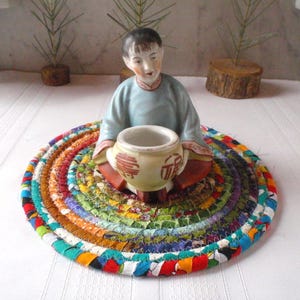 May include: A porcelain figurine of a person sitting on a colorful braided fabric coaster. The figurine is wearing a blue and white robe and holding a small pot.