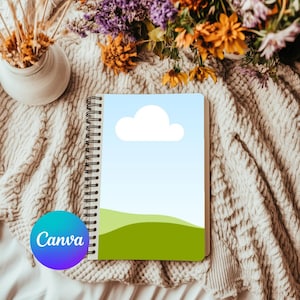 Pode incluir: Um caderno branco de espiral com uma ilustração de céu azul e grama verde na capa. O caderno tem uma nuvem branca no céu. O caderno está sobre um cobertor de malha branco.