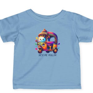 May include: A light blue t-shirt featuring a colorful illustration of an auto rickshaw with the text "Desi Me Rollin". The auto rickshaw is decorated with vibrant colors and has a smiling face.