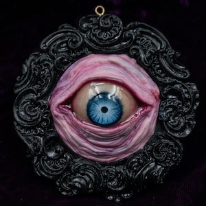May include: A unique, handcrafted pendant featuring a detailed, realistic eye with a vibrant blue iris. The eye is set within a black, ornate frame with swirling patterns. The eye is surrounded by pink and purple skin tones, creating a striking, surreal effect.