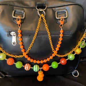 Puede incluir: Bolso de mano negro con detalles de cadenas de cuentas naranjas y amarillas. Las cadenas presentan varias cuentas de colores, incluyendo diseños naranjas, verdes y a rayas. El bolso tiene herrajes plateados y cierre de cremallera.