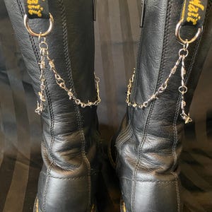 Puede incluir: Par de botas de cuero negro con costuras amarillas y una cadena plateada con detalles de alambre de púas. Las botas tienen cierre de cremallera y una etiqueta amarilla y negra que dice "AirWair".