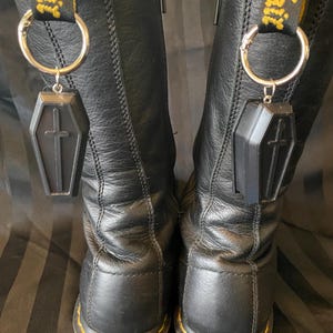 Puede incluir: Botas de cuero negro con colgantes en forma de ataúd colgando de las trabillas. Los colgantes son negros con un diseño de cruz. Las botas tienen costuras amarillas y el texto "Air Wair" en las trabillas.