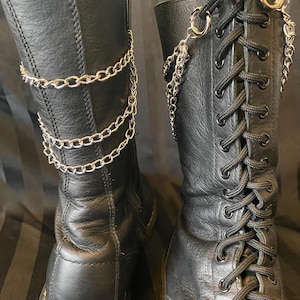 May include: Pair of black leather boots with silver chain detailing. One boot features a zip closure, the other has black laces. The boots have a classic design with yellow stitching around the soles.