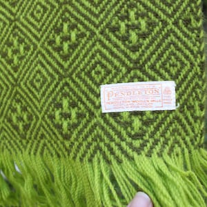 Könnte beinhalten: Nahaufnahme einer grün und olivgrün gewebten Decke mit einem geometrischen Rautenmuster. Die Decke hat einen Fransenrand an der Unterkante und ein rechteckiges Etikett mit Text. Das Etikett lautet "Pendleton Woolen Mills".