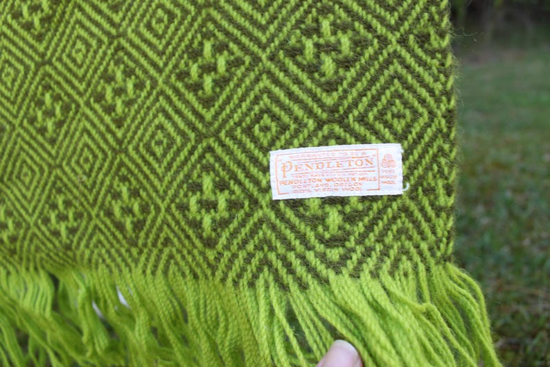 May include: Close-up of a green and olive-green woven blanket with a geometric diamond pattern. The blanket has a fringe along the bottom edge and a rectangular tag with text. The tag reads "Pendleton Woolen Mills".