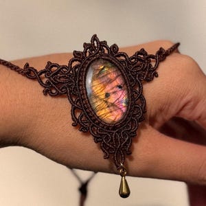 Könnte beinhalten: Ein dunkelbraunes Makramee-Armband mit einem ovalen Labradorit-Edelstein, der orangefarbene und violette irisierende Reflexe aufweist. Der Stein ist in aufwendige Knotenarbeit gefasst, mit einem kleinen, glatten, goldfarbenen Tropfenanhänger.