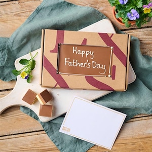 May include: A brown cardboard box with a window showing a chocolate bar with the words "Happy Father's Day" written in white icing. The box is sitting on a wooden surface with a green cloth and a small bouquet of flowers. There are also three small chocolate squares on a white chopping board.
