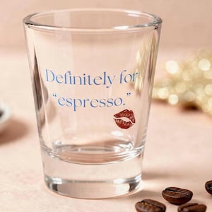 May include: Clear glass shot glass with the text "Definitely for 'espresso'" in blue and a red lip print. Coffee beans are scattered around the glass, suggesting a coffee-themed product.