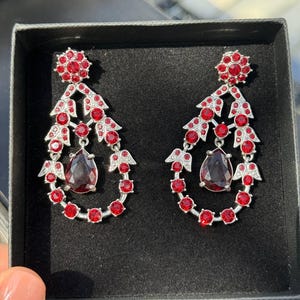 Op de afbeelding: Een paar sierlijke rode en zilveren oorbellen. Elke oorbel heeft een bloemmotief aan de bovenkant, een druppelvormige rode edelsteen en een reeks rode edelstenen. De oorbellen worden gepresenteerd in een zwarte doos.