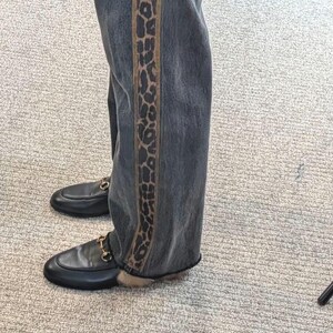 May include: Dark wash denim jeans with a leopard print side stripe. The jeans are paired with black leather loafers featuring gold-colored hardware.