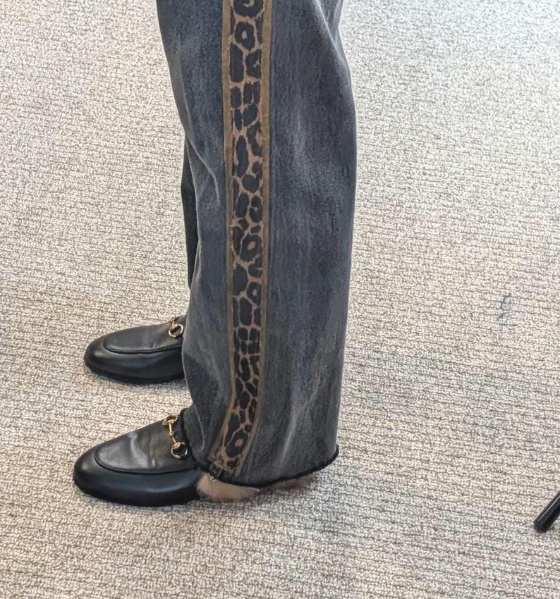 May include: Dark wash denim jeans with a leopard print side stripe. The jeans are paired with black leather loafers featuring gold-colored hardware.