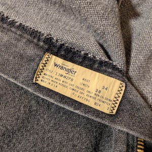 May include: Close-up of a pair of gray Wrangler jeans. The label reads "Wrangler" and includes the style number, waist size 38 inches, and length 34 inches. The jeans have a zipper and are made in the USA.