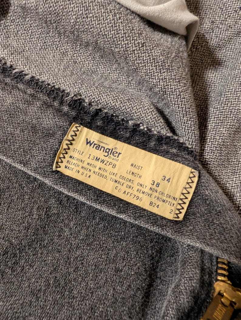 May include: Close-up of a pair of gray Wrangler jeans. The label reads "Wrangler" and includes the style number, waist size 38 inches, and length 34 inches. The jeans have a zipper and are made in the USA.