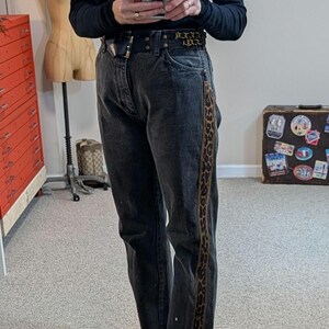 May include: Dark gray denim jeans with a leopard print stripe down the side. The jeans are worn with a black belt and black loafers with fur trim. The person is wearing a black long-sleeve shirt.