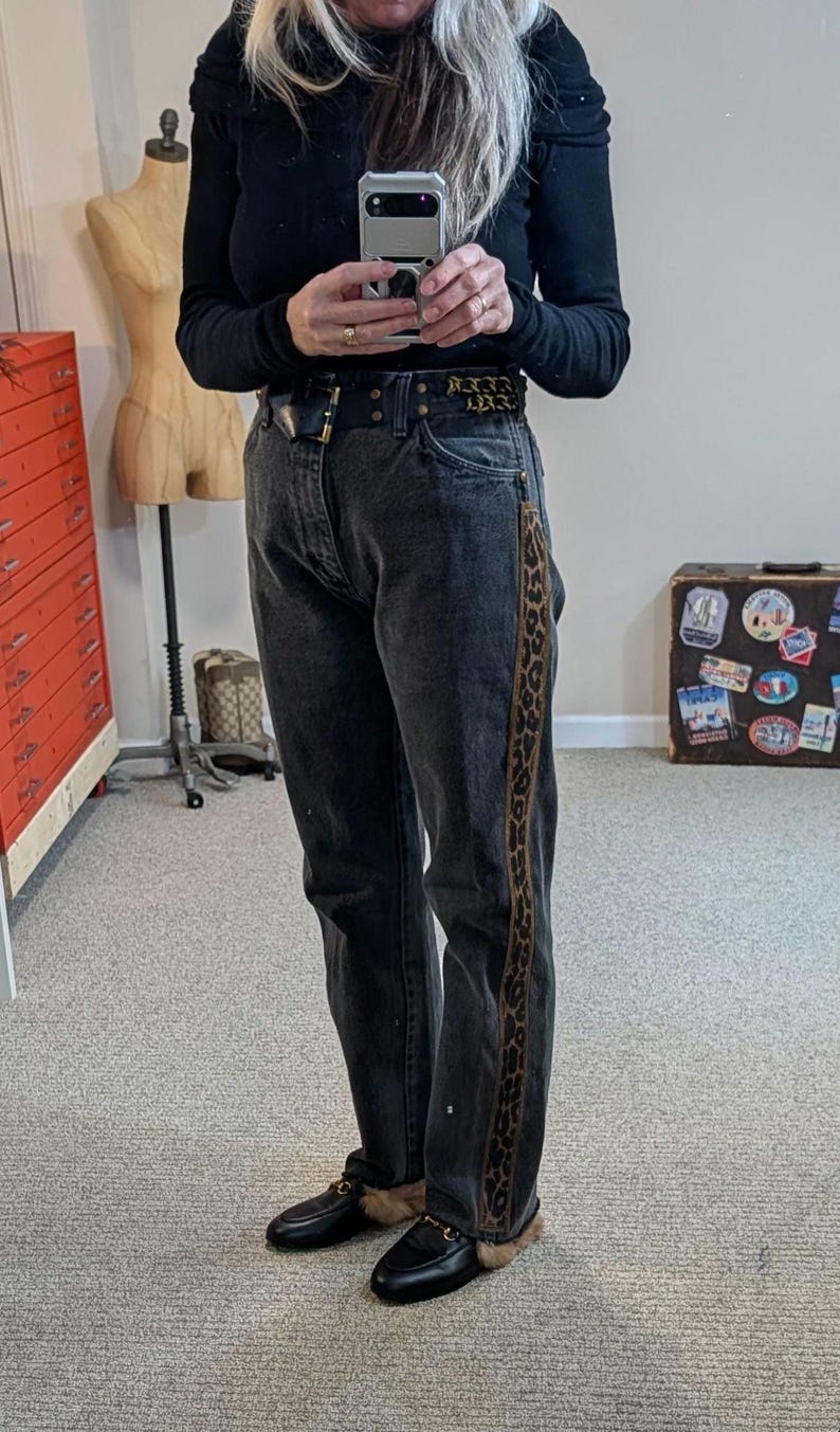 May include: Dark gray denim jeans with a leopard print stripe down the side. The jeans are worn with a black belt and black loafers with fur trim. The person is wearing a black long-sleeve shirt.