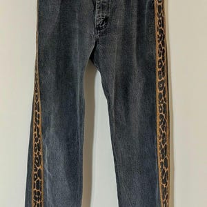 May include: Dark gray denim jeans with a straight leg cut. The jeans feature a vertical leopard print stripe down the outer seam of each leg. The jeans are hanging on a wooden hanger.