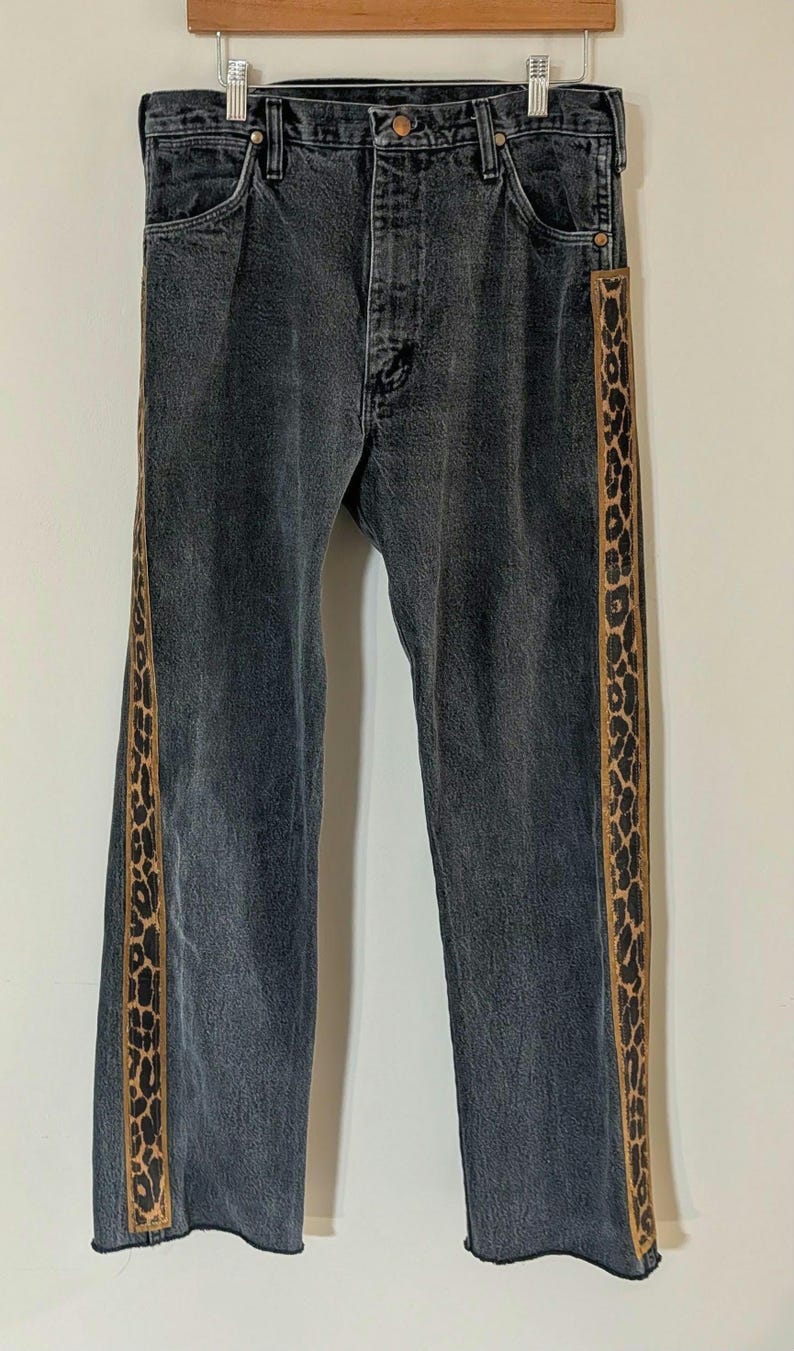 May include: Dark gray denim jeans with a straight leg cut. The jeans feature a vertical leopard print stripe down the outer seam of each leg. The jeans are hanging on a wooden hanger.