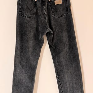 May include: Dark gray denim jeans with a classic five-pocket design. The jeans feature a straight leg cut and a leather patch on the back pocket. The Wrangler brand is visible on the patch.