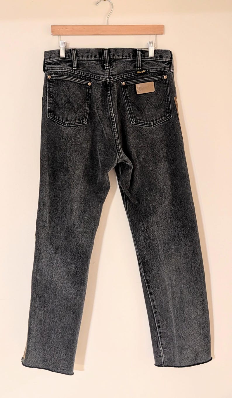 May include: Dark gray denim jeans with a classic five-pocket design. The jeans feature a straight leg cut and a leather patch on the back pocket. The Wrangler brand is visible on the patch.