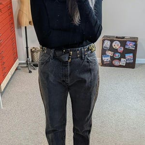 May include: A person wearing a black hat, black top, and dark wash jeans. The outfit includes a black belt with gold accents and black loafers with fur trim. A vintage suitcase with travel stickers is in the background.