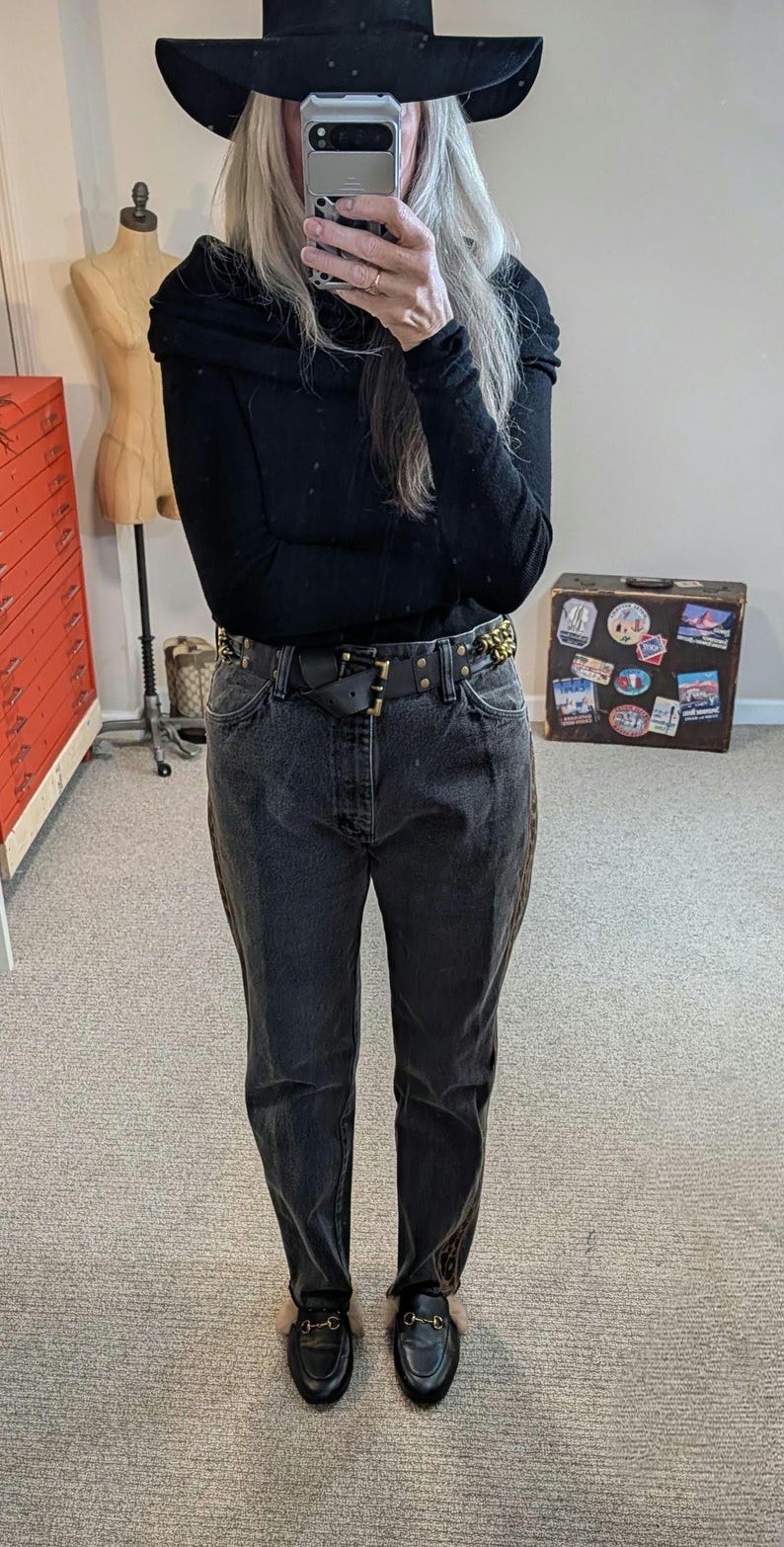 May include: A person wearing a black hat, black top, and dark wash jeans. The outfit includes a black belt with gold accents and black loafers with fur trim. A vintage suitcase with travel stickers is in the background.