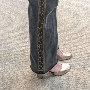May include: Dark wash denim jeans with a vertical leopard print stripe down the side. The jeans are paired with gold peep-toe heels. The outfit is casual and stylish.