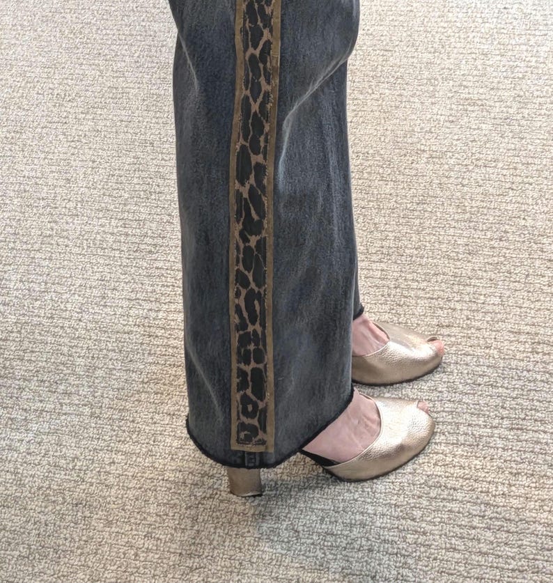 May include: Dark wash denim jeans with a vertical leopard print stripe down the side. The jeans are paired with gold peep-toe heels. The outfit is casual and stylish.