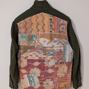 May include: A green utility jacket with a patchwork back panel made of colorful fabric scraps. The fabric scraps are sewn together in a random pattern and feature floral and geometric designs.