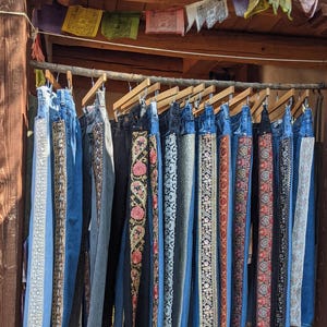 Könnte beinhalten: Eine Präsentation von Jeans aus Denim, die an Holzbügeln hängen. Die Jeans haben dekorative, bestickte Einsätze in verschiedenen Farben und Mustern an den Seiten. Die Jeans werden unter einem Holzbalken mit bunten Gebetsfahnen ausgestellt.