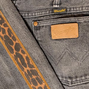 May include: Close-up of gray denim jeans with a brown and black leopard print stripe down the side. The back pocket features a brown leather patch with the Wrangler logo and a small tag with the Wrangler brand name.