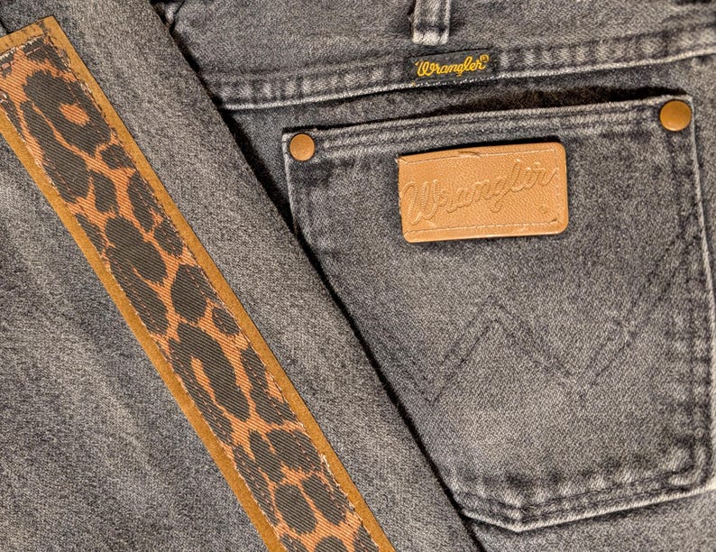 May include: Close-up of gray denim jeans with a brown and black leopard print stripe down the side. The back pocket features a brown leather patch with the Wrangler logo and a small tag with the Wrangler brand name.
