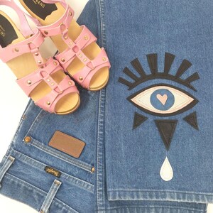 May include: Pink leather sandals with a wooden platform sole and gold studs. The sandals are paired with blue denim jeans with a black and white evil eye design and a white teardrop.