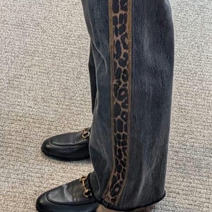 May include: Dark wash denim jeans with a leopard print stripe down the side. The jeans are paired with black leather loafers with gold-colored horsebit hardware.