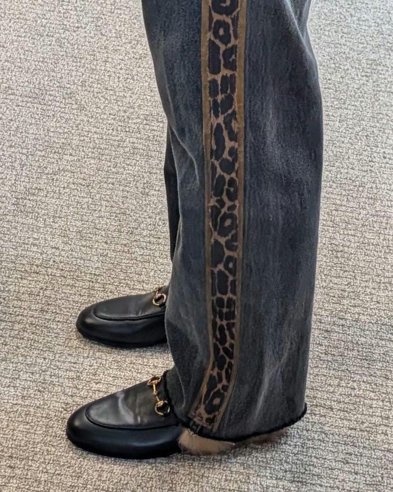 May include: Dark wash denim jeans with a leopard print stripe down the side. The jeans are paired with black leather loafers with gold-colored horsebit hardware.
