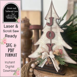 May include: A wooden Christmas tree decoration with the word "JOY" in red letters. The tree is painted white with a distressed finish and snowflake pattern. It is decorated with a bow, greenery, and a small tag. The text "Laser & Scroll Saw Ready" is visible.