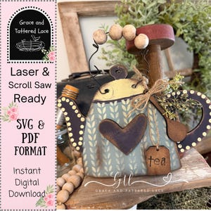 May include: A decorative wooden teapot with a distressed blue and yellow finish, featuring a heart-shaped accent and a small tag that says "tea." The teapot is adorned with wooden beads and a sprig of greenery. The text "Laser & Scroll Saw Ready" is visible.