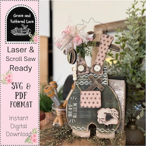 Op de afbeelding: Houten paasdecoratie in de vorm van een overall, groen geschilderd en versierd met roze stippen en een schaap. De tekst "HE IS RISEN" is zichtbaar. Versierd met een bloemenstrik en houten kralen. Klaar voor laser- en figuurzagen.