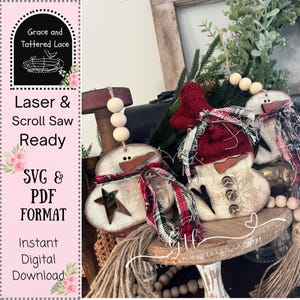 Pode incluir: Enfeites de boneco de neve de madeira com cachecóis xadrez vermelhos e pretos, narizes de cenoura e olhos de botão. Um tem um recorte de estrela. Os enfeites estão em uma bandeja de madeira com vegetação e um gorro vermelho.