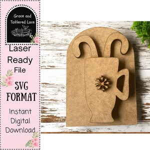 May include: A laser-ready file in SVG format for an instant digital download. The design features a wooden cutout of a mug with a snowflake and candy canes. The background is a light wood surface.
