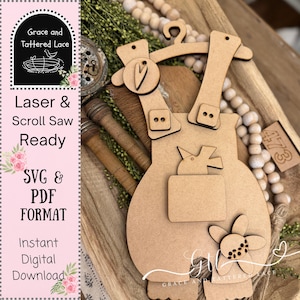 May include: A laser-cut wooden overalls design with bird and flower embellishments. The design includes the text "Laser & Scroll Saw Ready" and "SVG & PDF FORMAT". The overalls are displayed on a wooden tray with decorative beads and greenery.