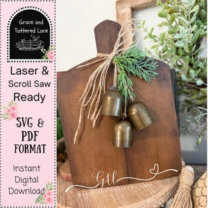 May include: A rustic, brown wooden cutting board with a decorative arrangement. It features three small, antique-style bells, tied with twine and greenery. The board has a handle and is decorated with the text "Grace and Tattered Lace", and "Laser & Scroll Saw Ready".