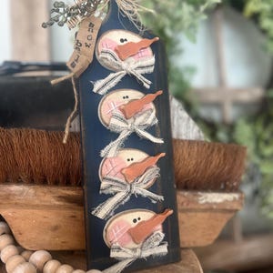 Op de afbeelding: Een marineblauwe houten sneeuwmanversiering met vier sneeuwmannengezichten. Elk gezicht heeft een roze wang, een oranje wortelneus en een zwart-wit lintstrik. De decoratie heeft een label met de tekst "Snow Biz".