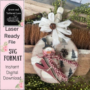 Op de afbeelding: Rustieke houten kerstversiering met een sneeuwpop met ski's, een hoge hoed en een rood-witte sjaal. De versiering heeft een verweerde afwerking en is versierd met een sneeuwvlok en kleine bomen. De tekst "Grace and Tattered Lace" is zichtbaar.