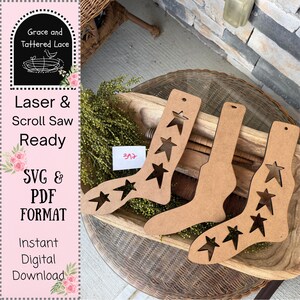 May include: Three brown wooden sock cutouts with star cutouts. The socks are displayed on a wooden tray with greenery. The image also includes a pink and white graphic with the text "Grace and Tattered Lace", "Laser & Scroll Saw Ready", "SVG & PDF FORMAT", and "Instant Digital Download".