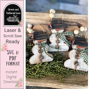 Peut inclure: Ornements de bonhomme de neige en bois avec des chapeaux noirs, des nez de carotte et des écharpes à carreaux verts. Chaque bonhomme de neige a des boutons en bois et le mot "brrr". Les ornements sont exposés sur un plateau en bois avec de la verdure.