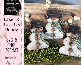 May include: Wooden snowman ornaments with black hats, carrot noses, and green plaid scarves. Each snowman has wooden buttons and the word "brrr" on the bottom. The ornaments are displayed on a wooden tray with greenery.