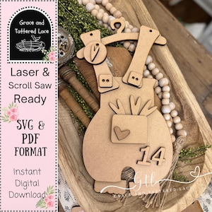 Puede incluir: Una forma de peto de bebé de madera cortada con láser con el número 14. El peto tiene un bolsillo con un recorte de corazón y se muestra en una bandeja de madera con cuentas decorativas y vegetación. El texto "Laser & Scroll Saw Ready" es visible.